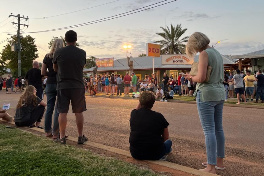 People stand in the street of a small outback town under a bright light blue sky where the sun is setting