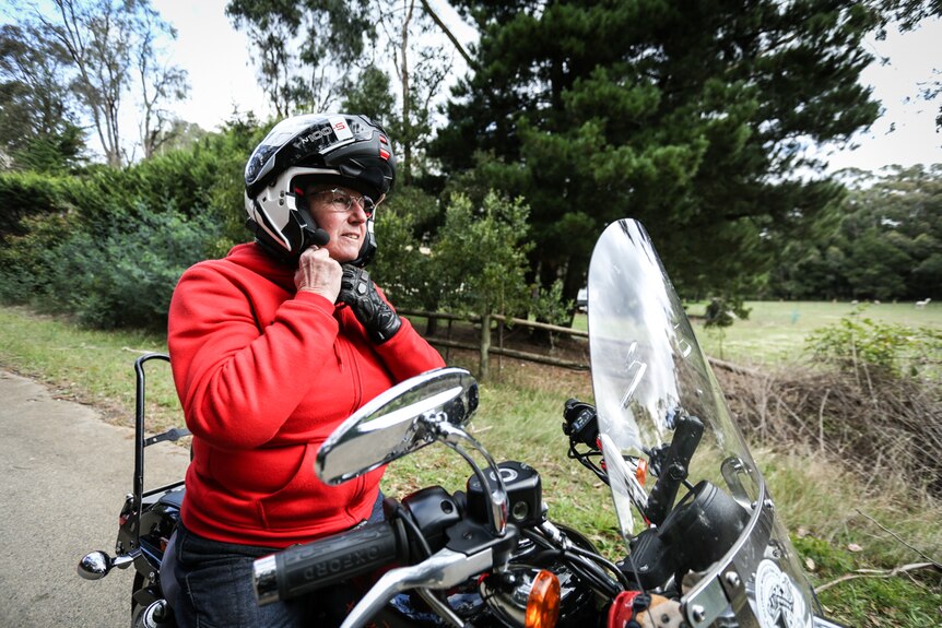 A woman wearing a red hoodie sits astride a motorcycle. She is fastening the chinstrap on her helmet.