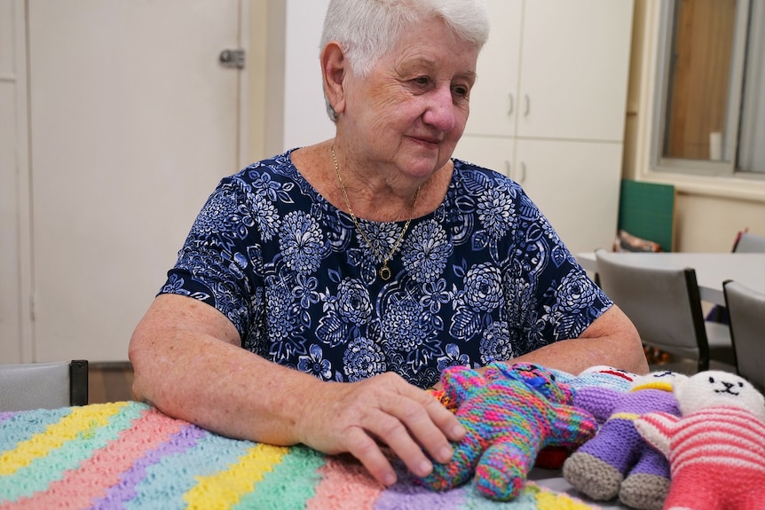 Jan Rawlings, blue floral shirt, grey hair sitting. Her hands touch some colourful, crocheted trauma teddies, slight smile.