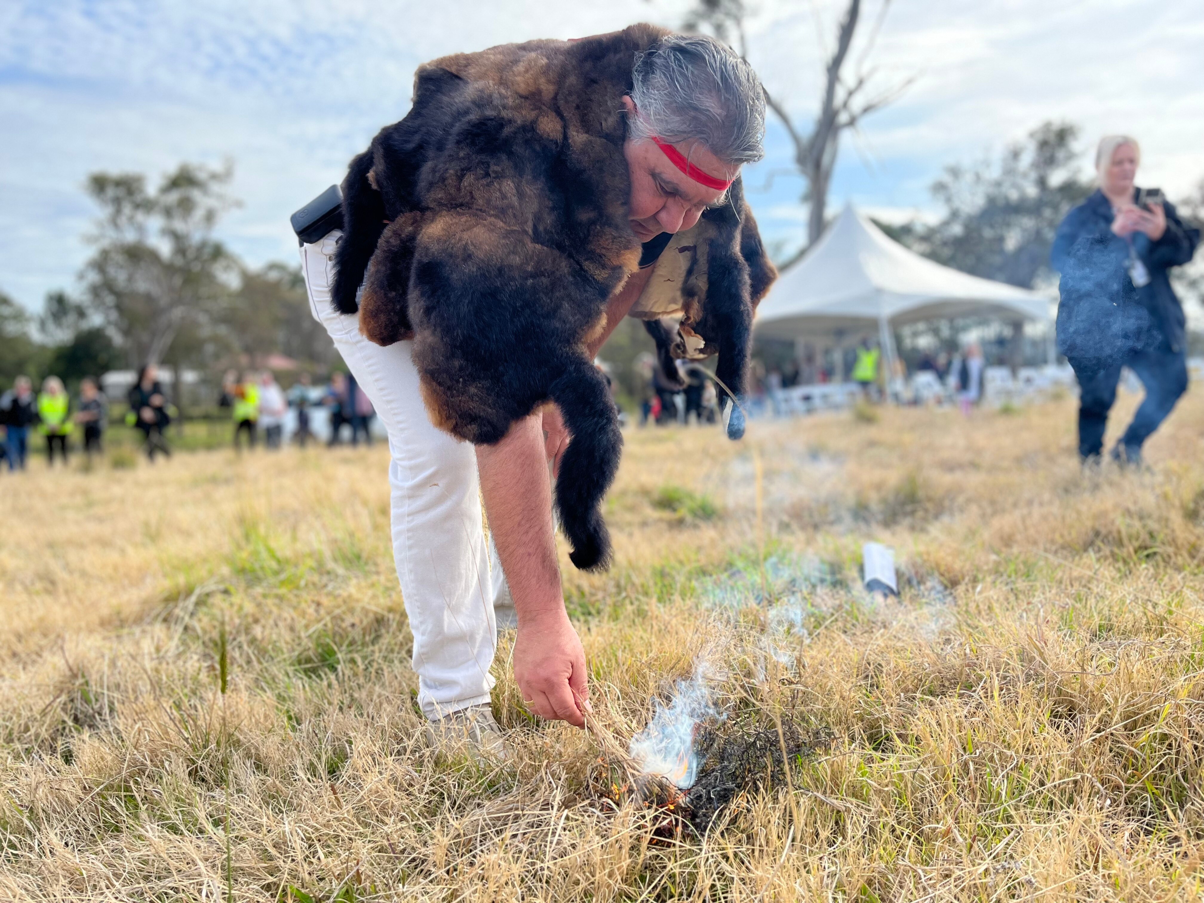 An Indigenous man wearing a possum cloak lights some grass on fire.