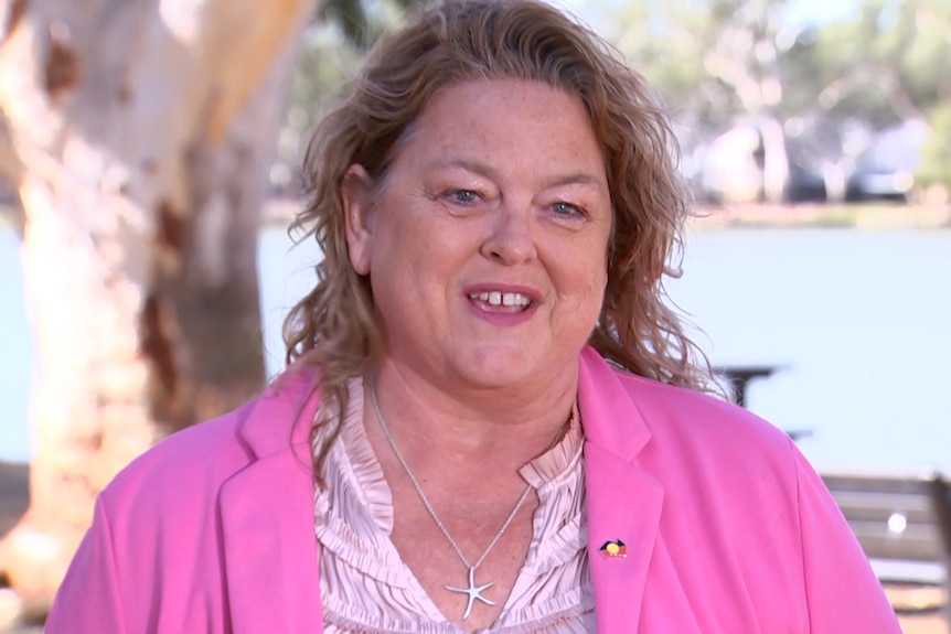 A woman wearing a pink blazer and slightly smiling stands in front of a river