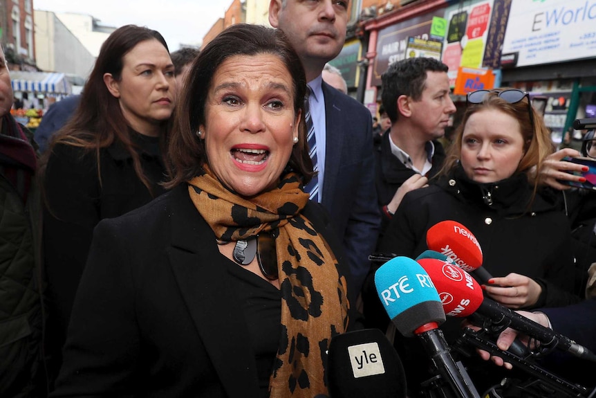 A woman in a bronze and black patterned scarf speaks surrounded by reporters with microphones.