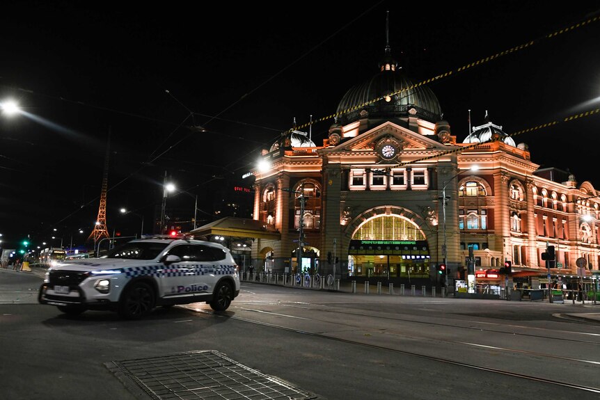 A white police car turns in front of a well-lit brown building at night on a deserted city street.