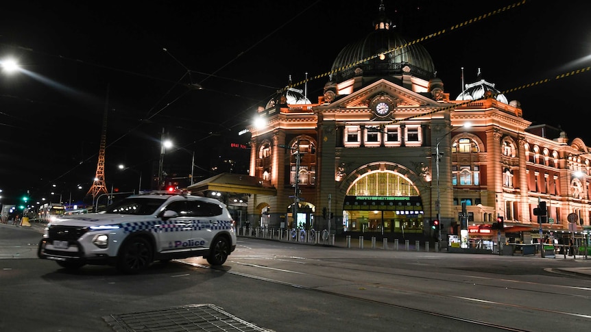 A white police car turns in front of a well-lit brown building at night on a deserted city street.