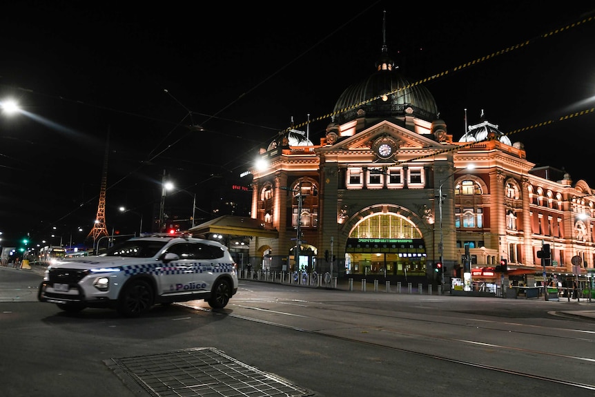 A white police car turns in front of a well-lit brown building at night on a deserted city street.