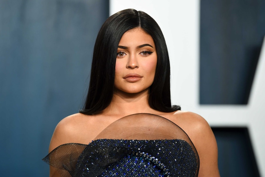 Kylie Jenner looks slightly to the right as she poses for photographs against a blue and white backdrop in a blue and black gown