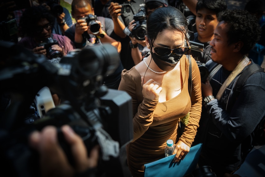 A woman wearing sunglasses, a dark face mask and brown dress makes her way through a press pack with cameras