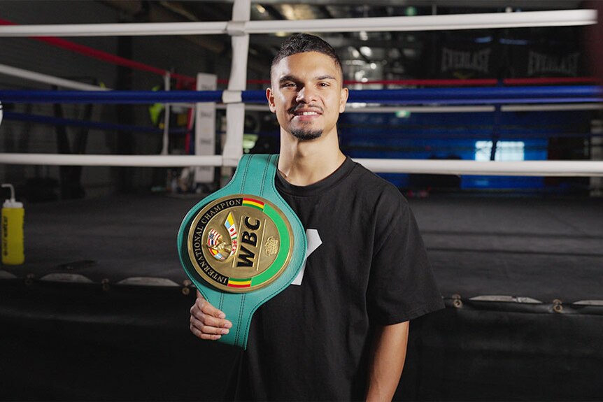 A young Aboriginal man with a boxing belt over his shoulder smiles at the camera in front of a boxing ring.
