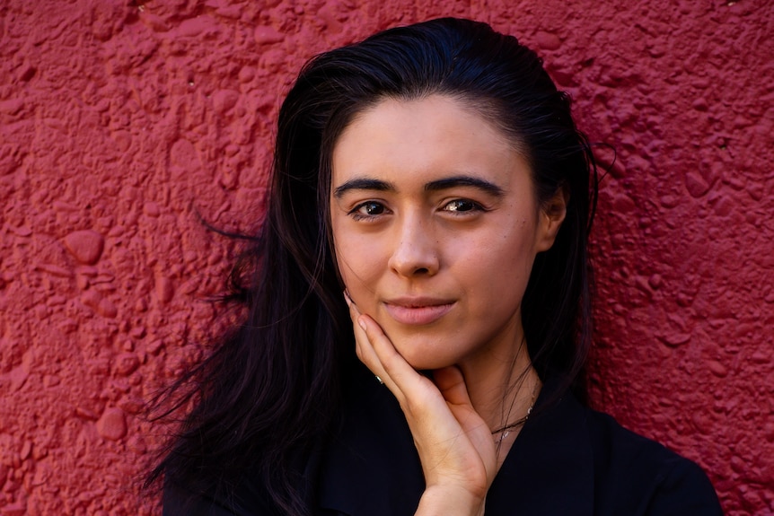 A young Tongan-Australian woman with long dark hair standing in front of a red wall, with her hand on her chin