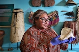 Fijian Tongan woman stands holding a shell wearing latex gloves with historic pacific clothes hung behind her
