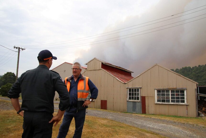 Phil Vickers stands in front of a shed-like structure, there's a smoke plume in the background.