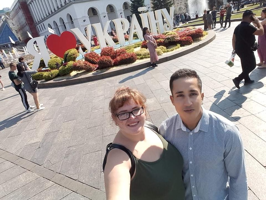 A couple pose for a selfie in a city square with a large white sign in reverse with a heart set in a garden as people mingle.