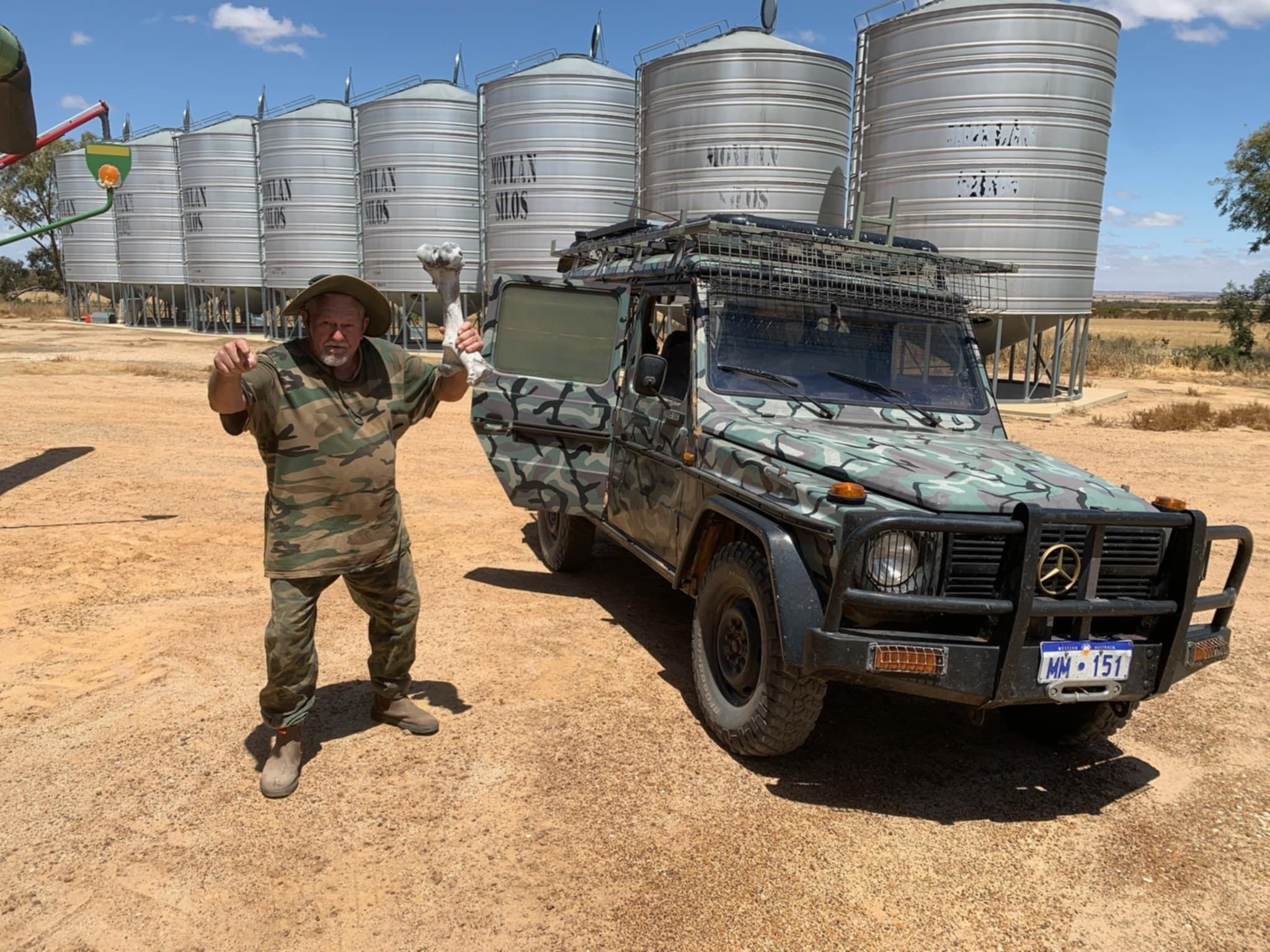 Um homem baixo vestido de camuflagem, parado ao lado de um carro camuflado, segura um grande osso em uma pose boba, perto dos silos.
