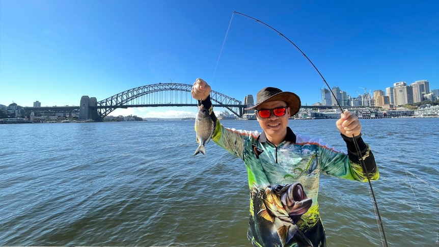 Chi Lam with a fishing rod and fish in front of Sydney Harbour Bridge.
