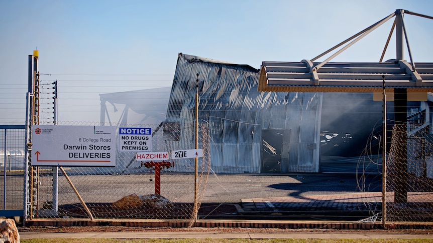 A fire-damaged corrugated iron warehouse, surrounded by a car park and chain fence.