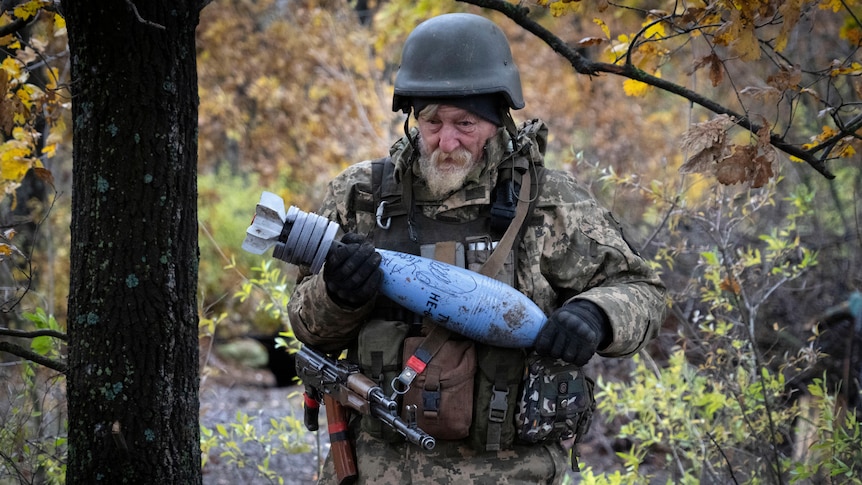 A soldier in the bush holds a blue shell with hand writing on it.