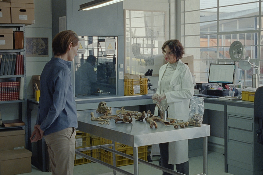 A middle-aged woman stands at a table of human bones in a scientific laboratory across with another woman in a lab coat