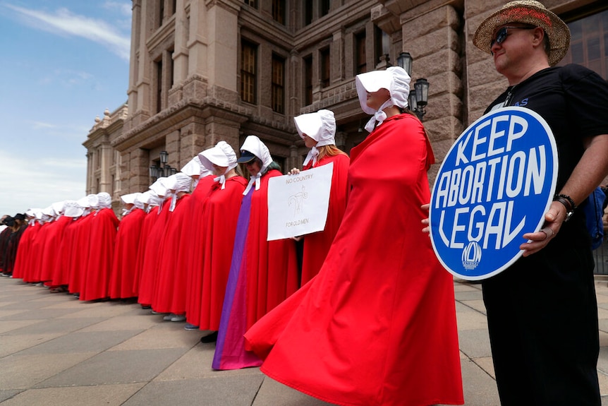 Women in white bonnets and red robes stand in front of a government building next to a man holding a Keep Abortion Legal sign.
