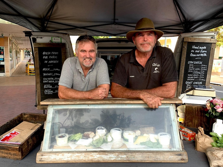 Two smiling, bearded men lean on a display cabinet at a farmers' market