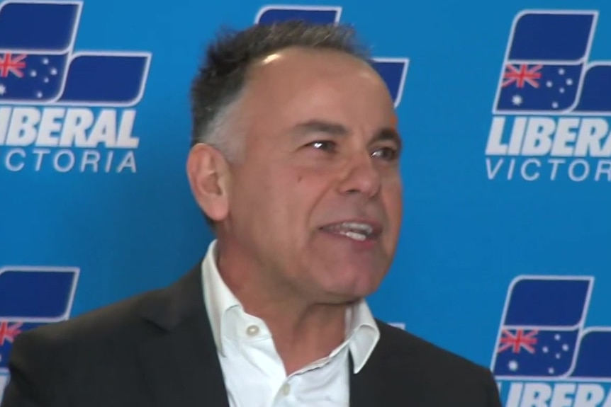 John Pesutto smiles, holding a microphone in front of a Liberal Party backdrop.