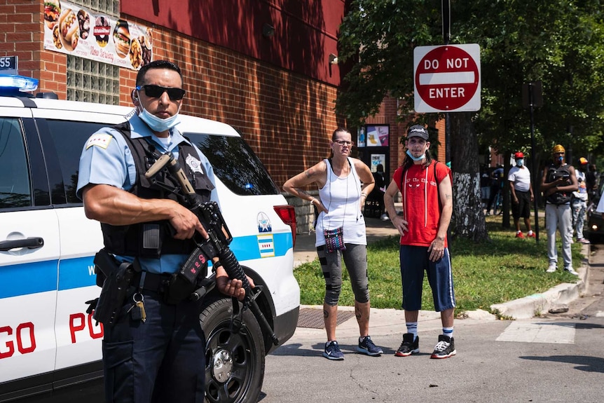 A police officer with a large gun and a face mask stands to the left, with a number of people looking on