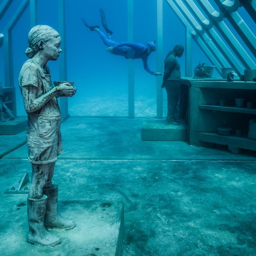 A diver swims in a metal underwater structure at the Museum of Underwater Art.  Inside there are statues of a girl and a woman.