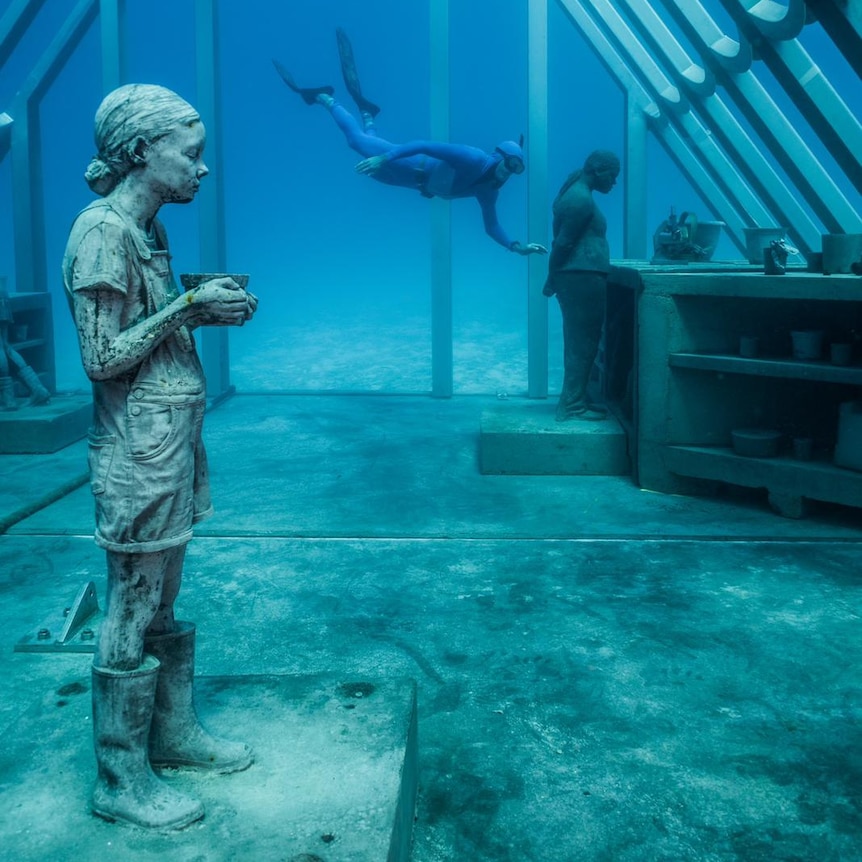 A diver swims into a metal underwater structure at the Museum of Underwater Art. Inside there are statues of a girl and a woman.