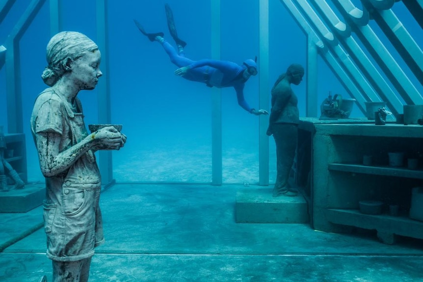 A diver swims into a metal underwater structure housing statues of a girl and a woman.
