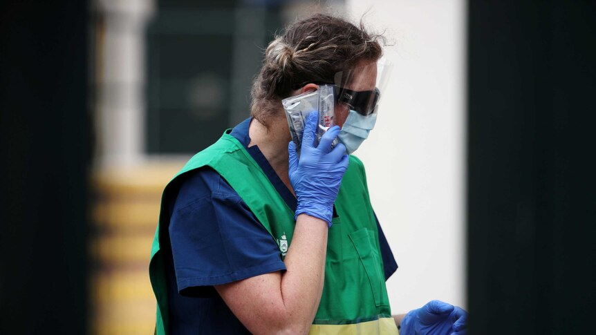 A healthcare worker in gloves, a face mask and a green bib speaks on her mobile phone which is wrapped in plastic