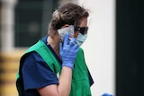 A healthcare worker in gloves, a face mask and a green bib speaks on her mobile phone which is wrapped in plastic
