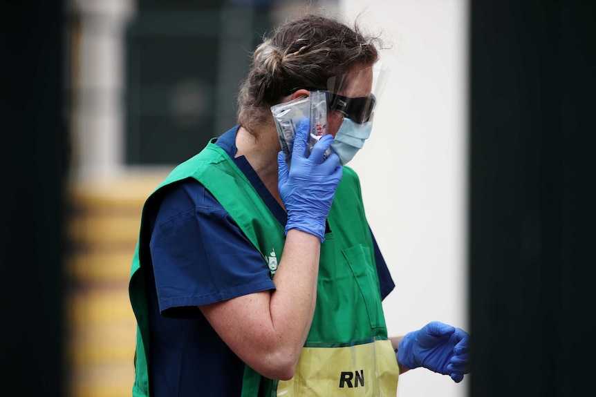 A healthcare worker in gloves, a face mask and a green bib speaks on her mobile phone which is wrapped in plastic