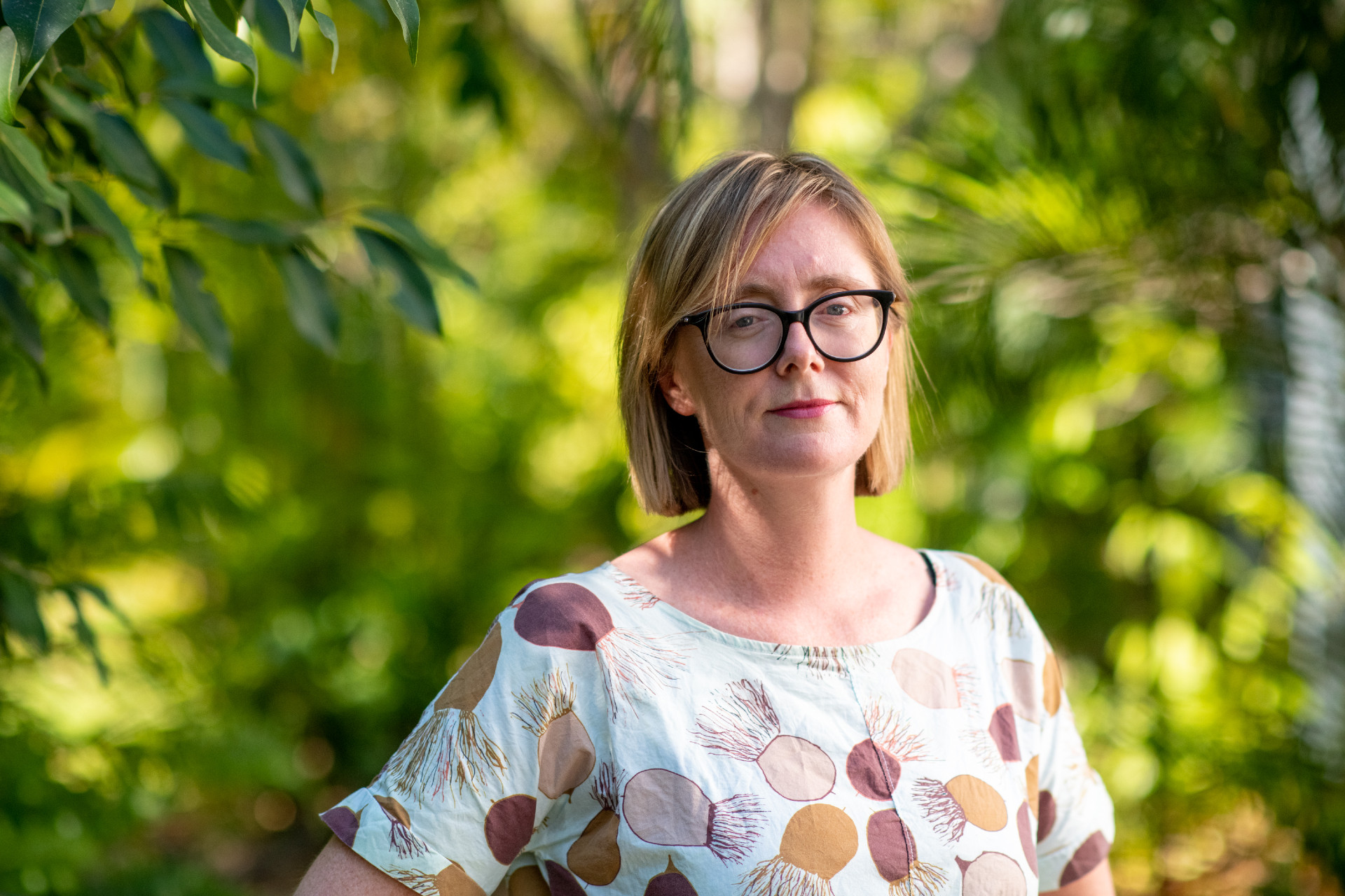 A woman wearing glasses stands in a leafy Darwin backyard, looking slightly concerned.