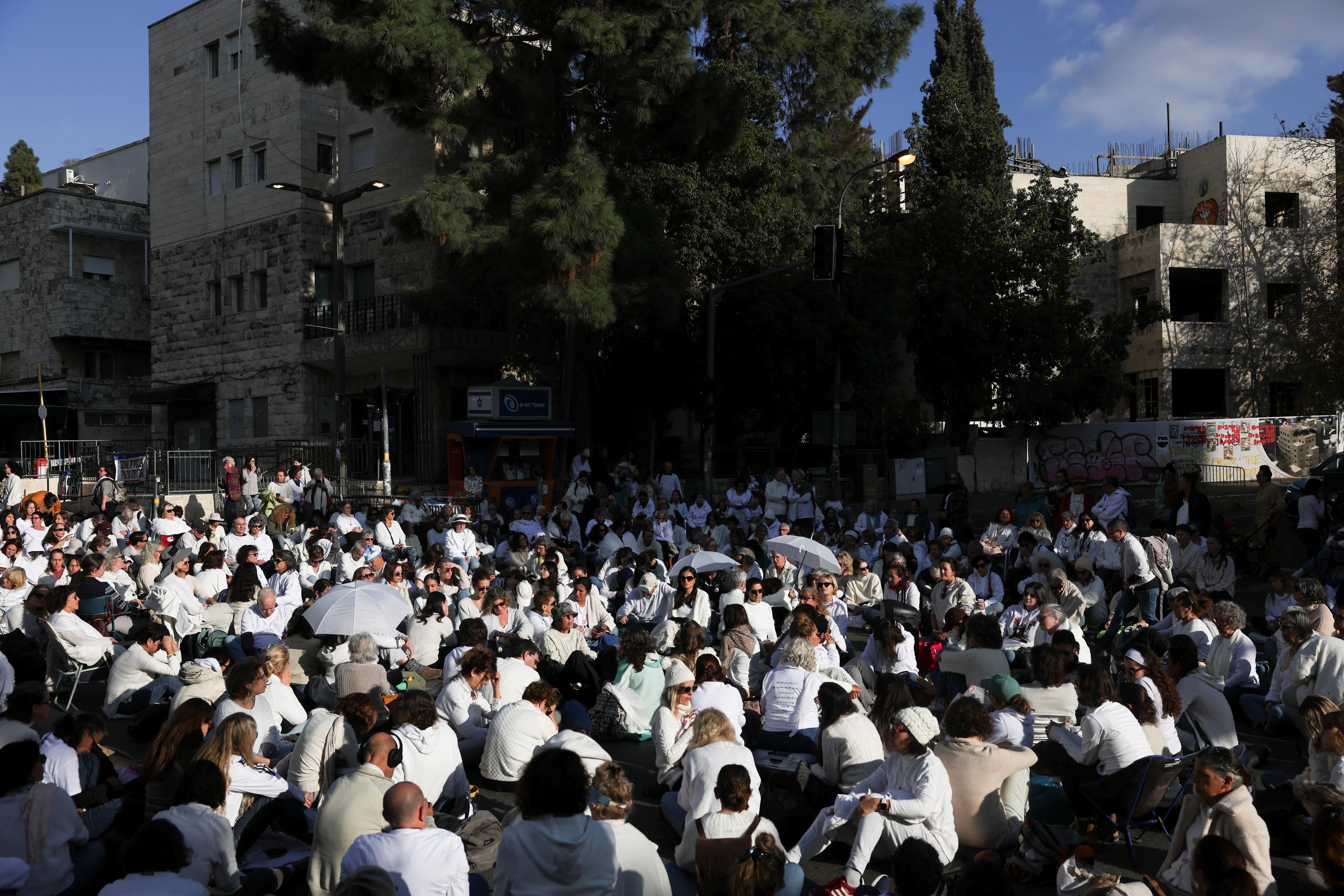 A crowd of protesters all wearing white clothing kneeling or sitting in front of a building and dark silhouette of a tree
