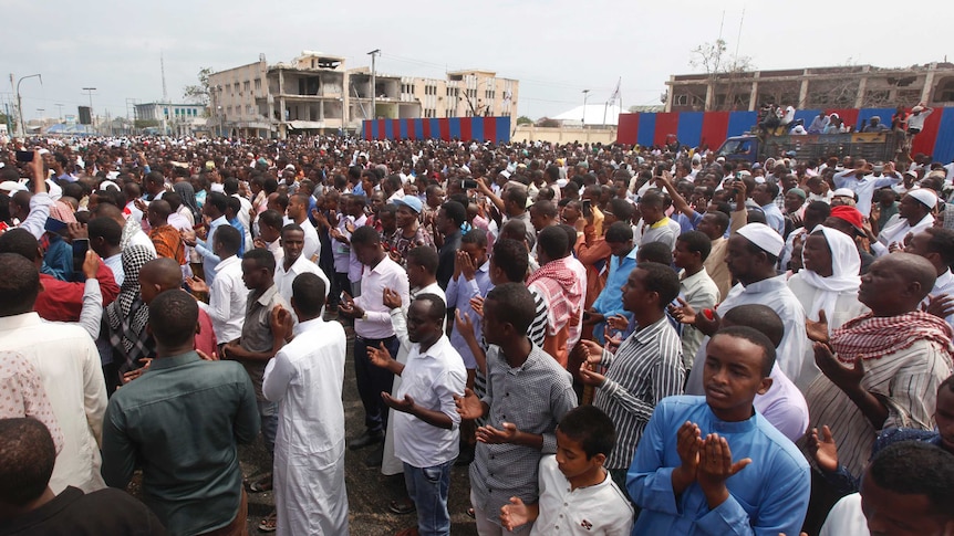 A huge crowd of people gather in front of destroyed buildings.