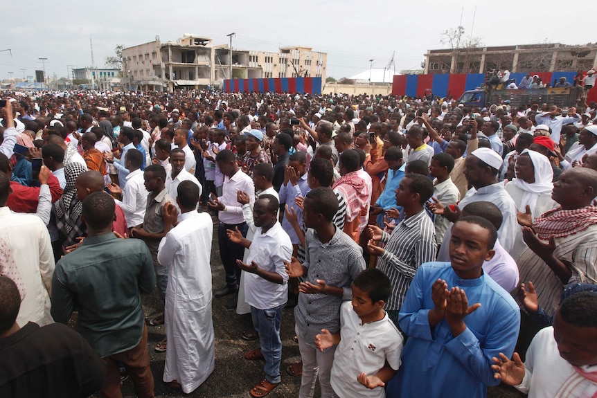 A huge crowd of people gather in front of destroyed buildings.