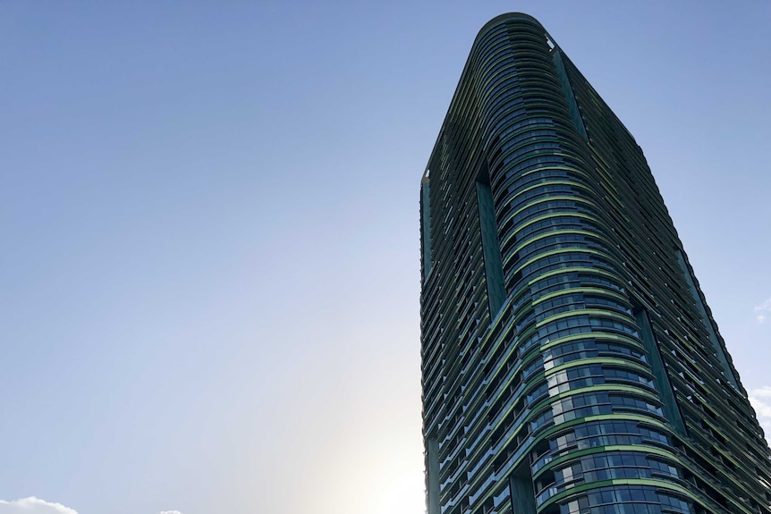 A blue and green high-rise building is seen to the right of frame with clear blue skies surrounding the skyscraper.