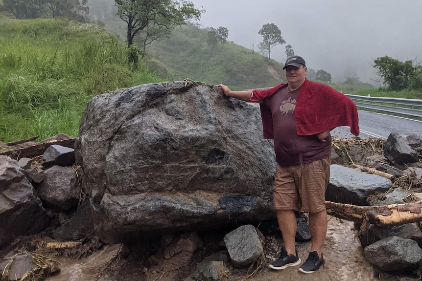 A man standing next to a large boulder that's fallen onto the road in a landslip.