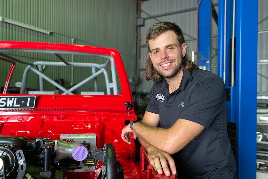 A man with a short, brown mullet haircut leans on a bright red car with an exposed engine