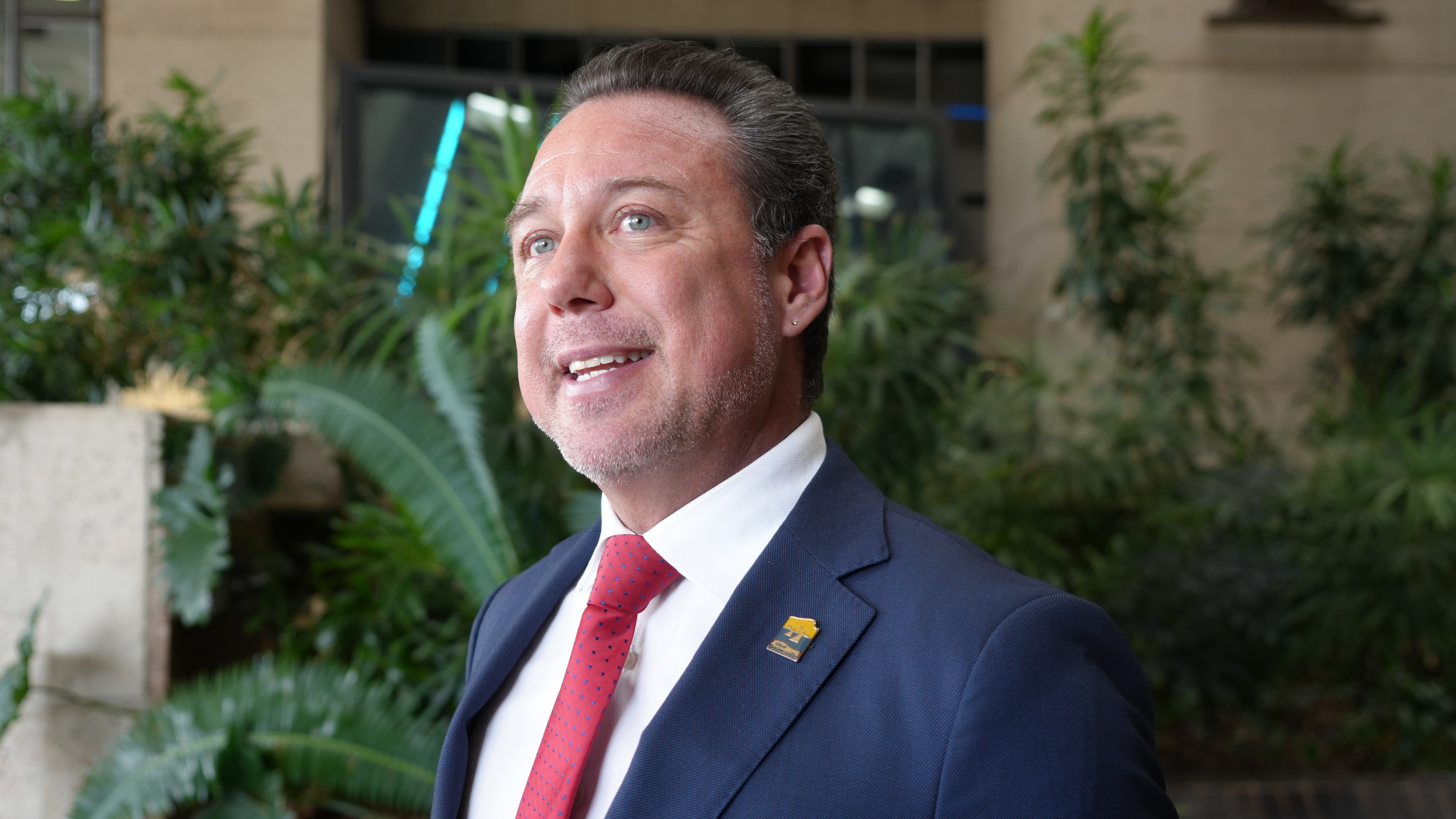 A middle aged man with greying pushed back hair in a blue suit and red tie.