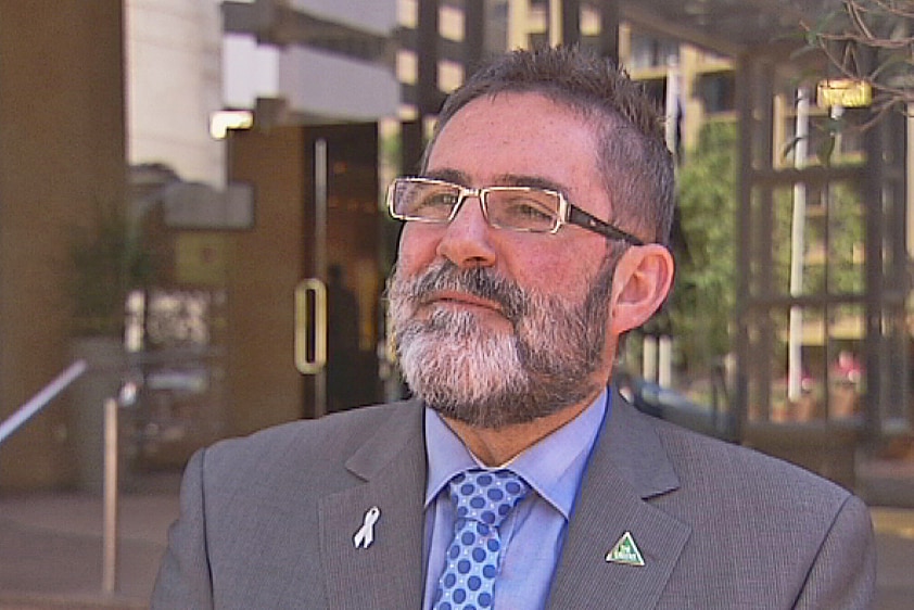 A white man wearing a suit and glasses. His hair and beard are black and grey