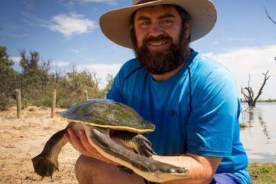 A man stands on the side of a body of water holding a large turtle