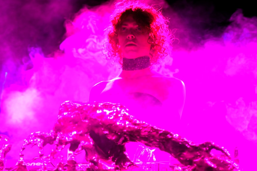 A performer with short hair stands in front of a smoke screen surround in pink light.
