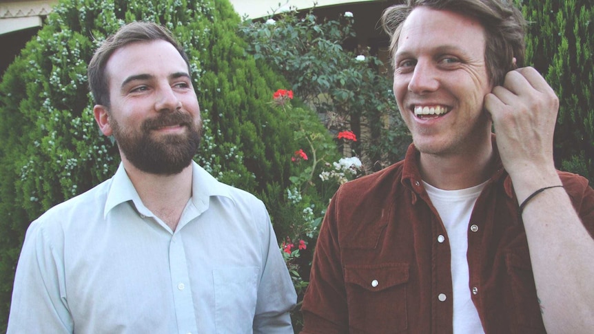 Two men standing side by side and smiling, in front of a garden.