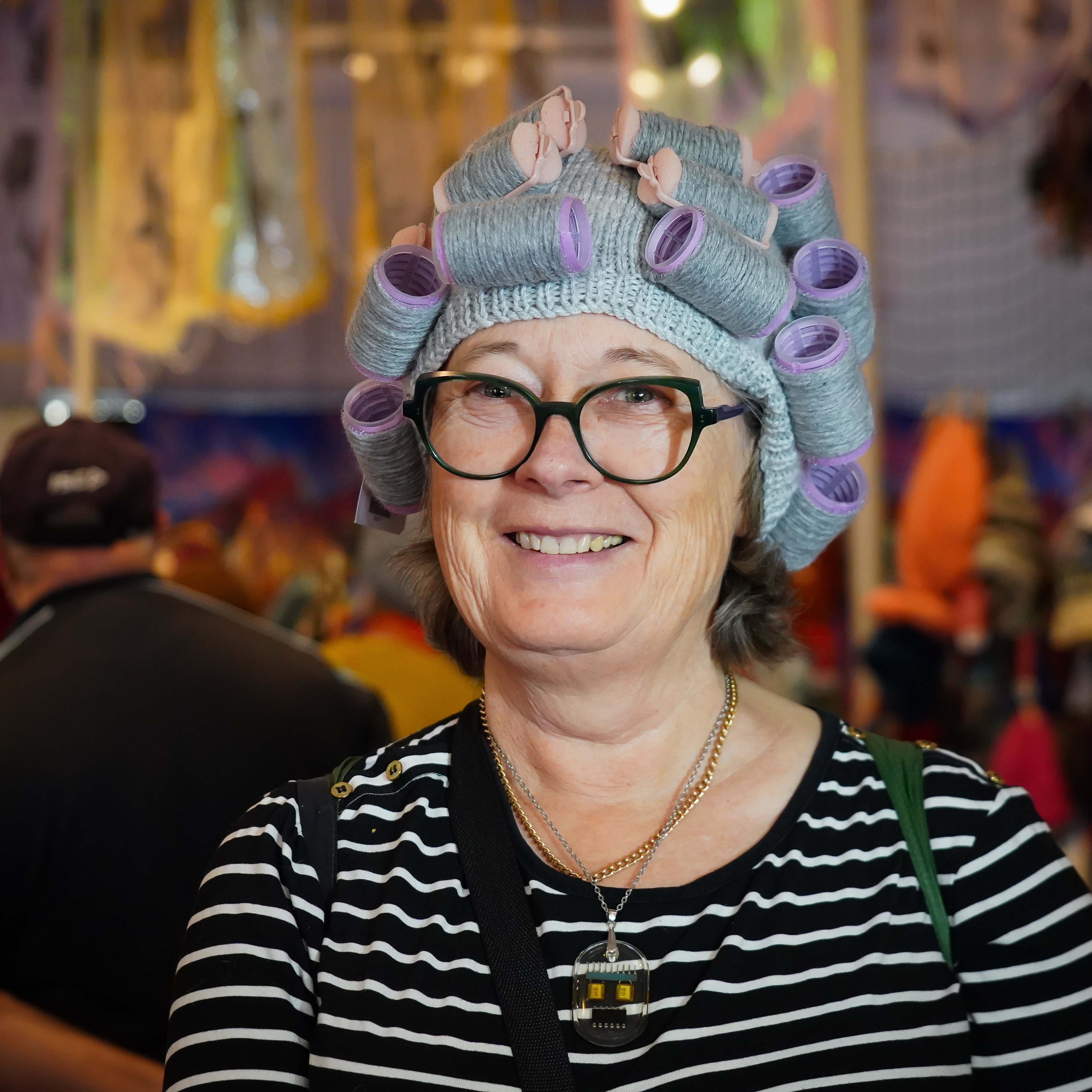 A woman wearing reading glasses and a grey beanie, pink and purple hair rollers are attached to her knitted hat.