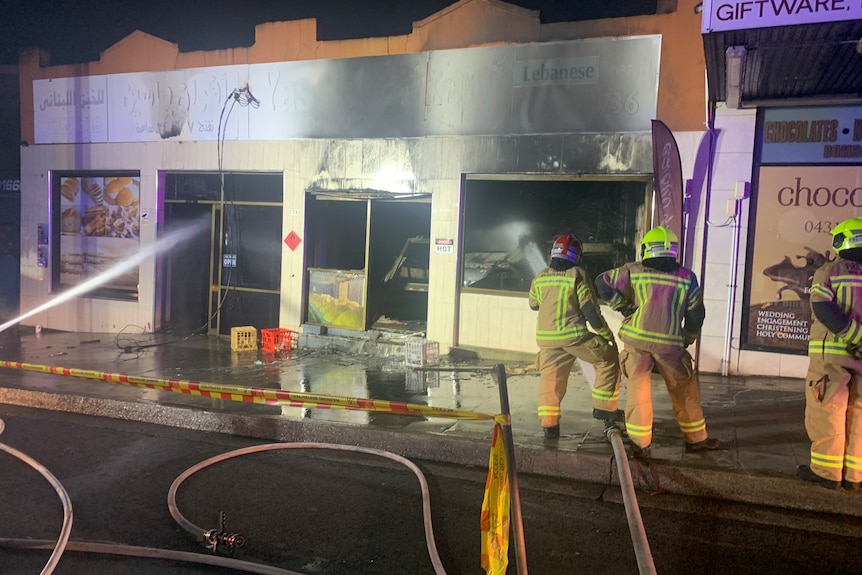 External of small shop front that appears to have been gutted by fier. Two people stand on the right in yellow fire uniform