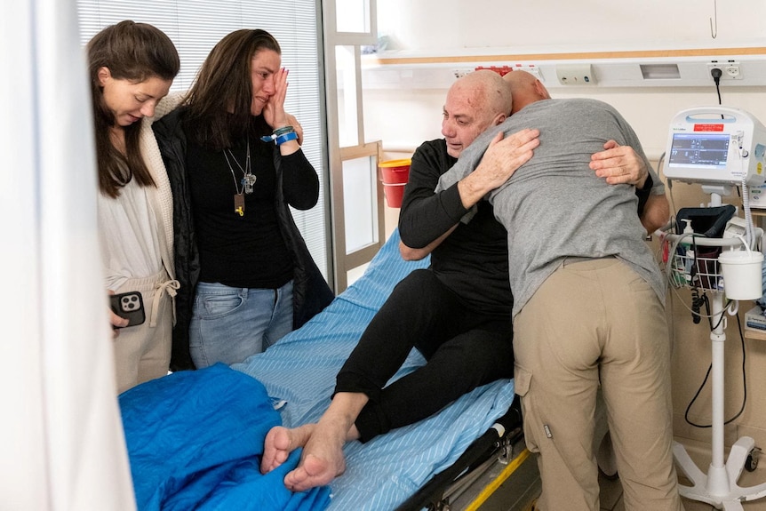 a man in black shirt and pants on a hispital is hugged by a  man as two women stand by bed crying