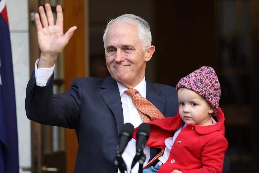Malcolm Turnbull waves and smiles while holding a young child, australian flag in background.