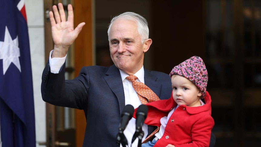 Malcolm Turnbull waves and smiles while holding a young child, australian flag in background.