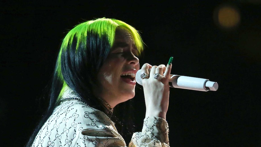 A woman with green hair sings in darkness on stage