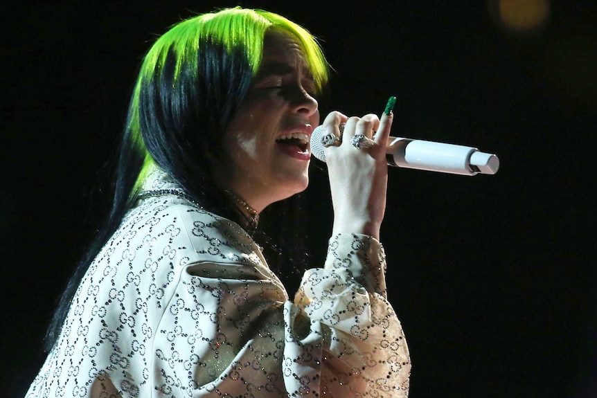 A woman with green hair sings in darkness on stage